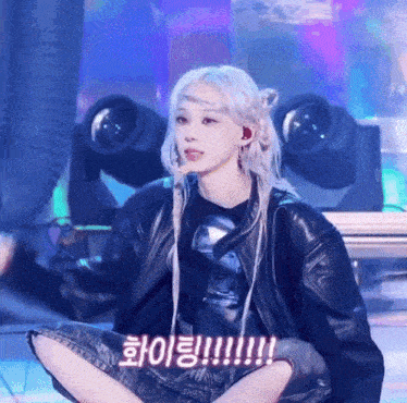 a woman in a black jacket sits on the floor with her legs crossed in front of a bunch of stage lights