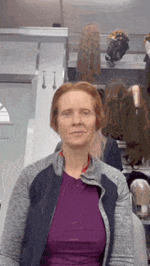a woman wearing a purple shirt and a gray jacket is standing in front of a shelf with wigs .