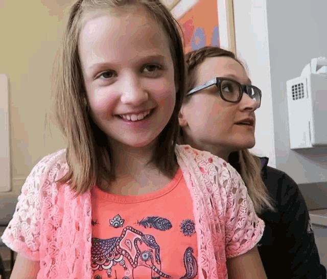 a little girl wearing a pink shirt with an elephant on it smiles with a woman wearing glasses behind her