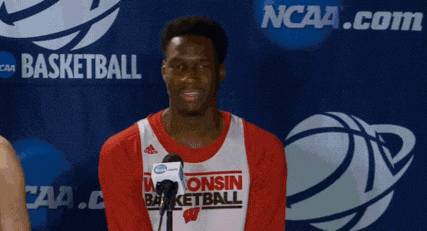 a man wearing a consin basketball jersey stands in front of a ncaa logo