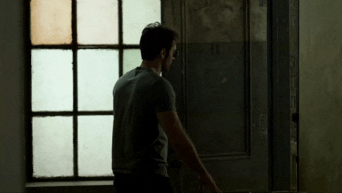 a man in a grey shirt is standing in front of a window looking out