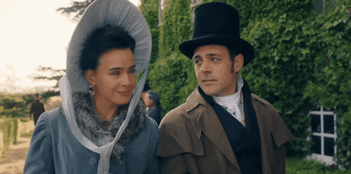 a man and a woman are standing next to each other in a garden . the man is wearing a top hat .