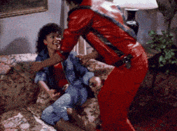 a man in a red leather jacket stands next to a woman on a couch