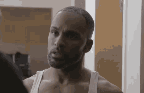 a man with a beard and a white tank top is looking at himself in the mirror .