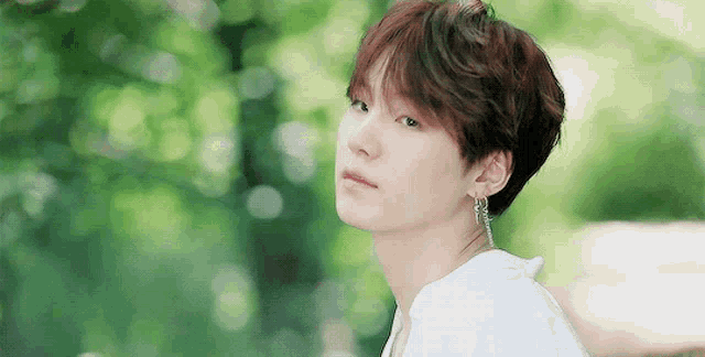 a young man wearing a white shirt and earrings is standing in front of trees .