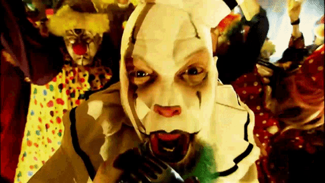 a clown in a white mask is holding a knife in his mouth