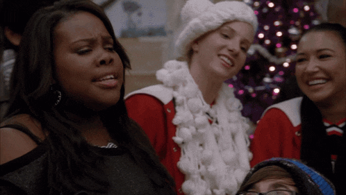 a group of girls are singing in front of a christmas tree and one girl is wearing a santa hat