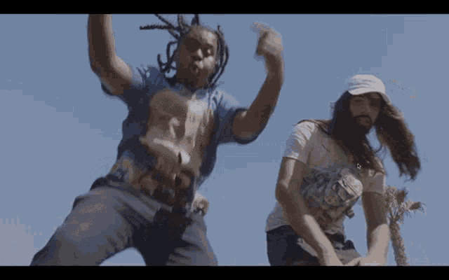 two men are dancing in front of a blue sky . one of the men has dreadlocks and the other has a beard .