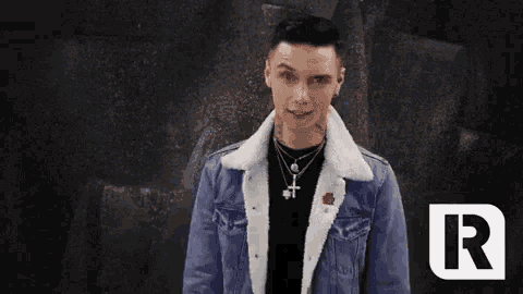 a man wearing a denim jacket and a necklace is standing in front of a wall .