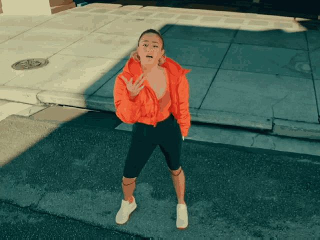 a woman wearing an orange jacket and black shorts is standing on a sidewalk