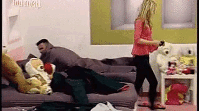 a woman is standing next to a man laying on a couch with a teddy bear .