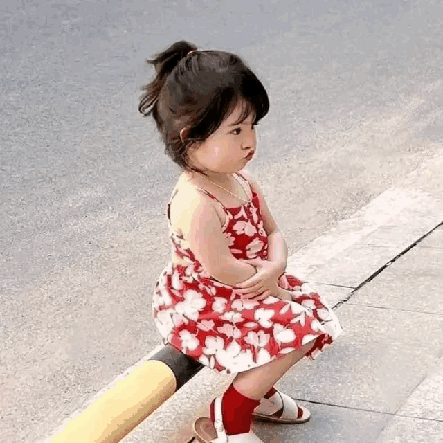 a little girl in a red and white dress is sitting on the sidewalk .