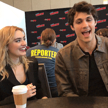 a man and a woman sit at a table with a woman wearing a reporter shirt