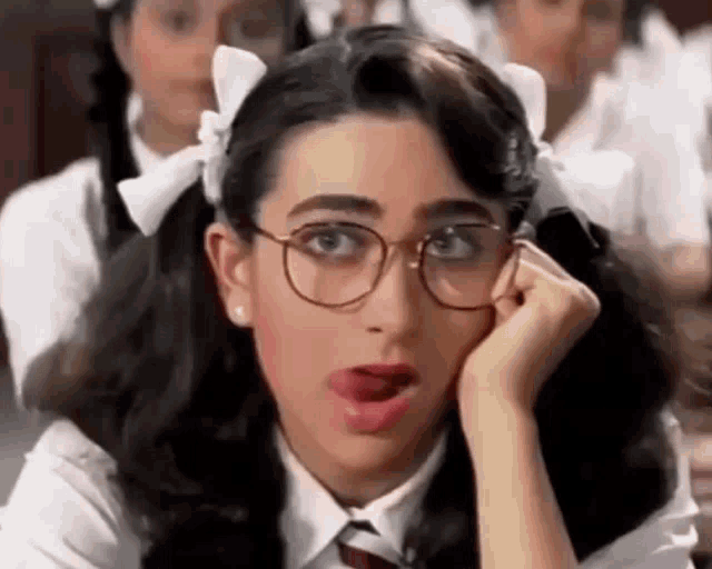 a girl wearing glasses and pigtails is sitting in a classroom with her hand on her face .