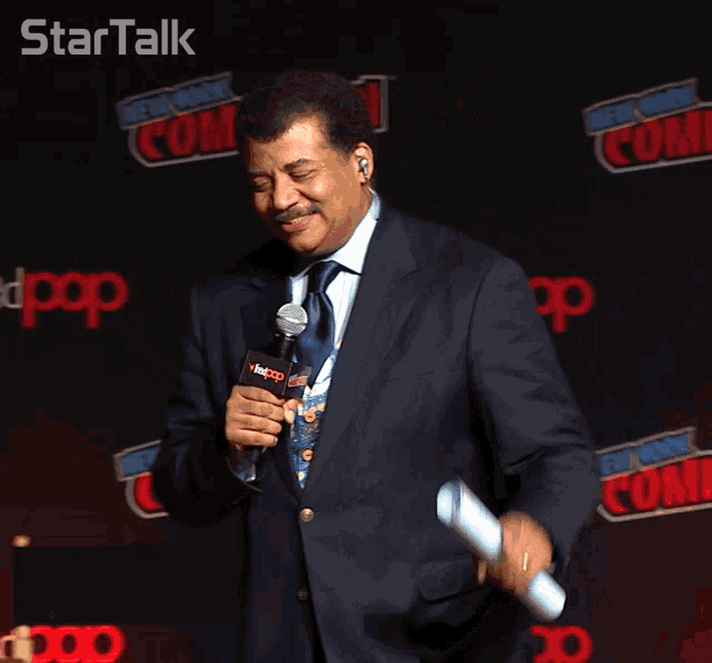 a man in a suit and tie holds a microphone in front of a new york comic con banner