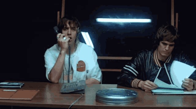 two men are sitting at a table with one talking on a phone and the other looking at a piece of paper