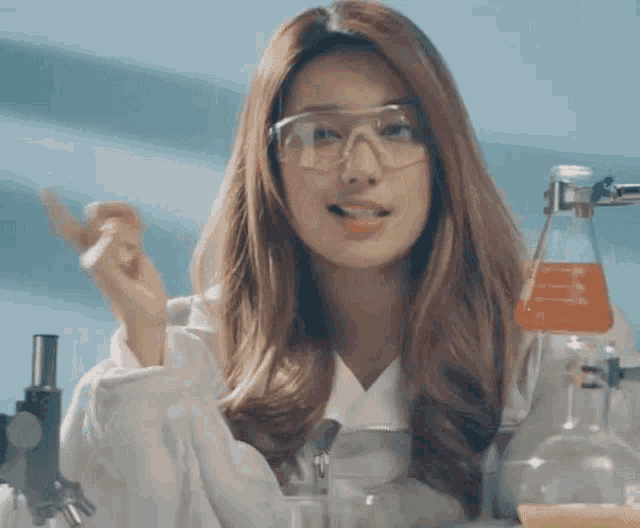 a woman wearing glasses and a lab coat is standing in front of a beaker with orange liquid in it .