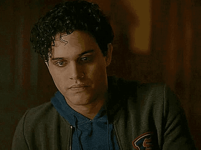 a young man with curly hair is wearing a gray jacket and a blue sweater and looking at the camera .