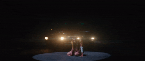a woman is kneeling on the ground in front of a car with the lights on