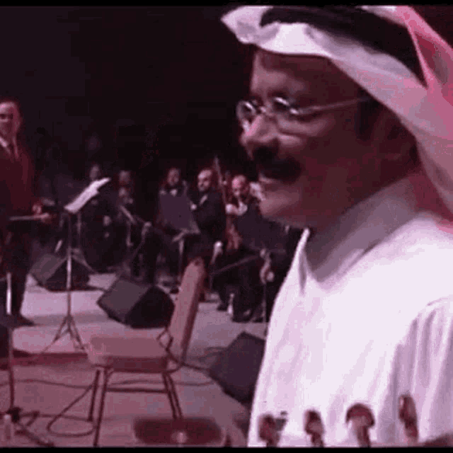 a man with glasses and a mustache is standing on a stage with an orchestra in the background .