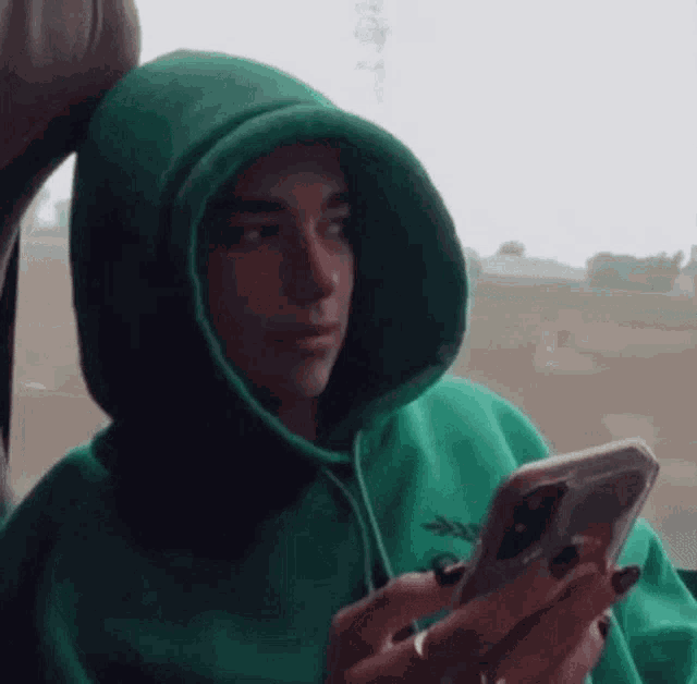 a person wearing a green hoodie is looking at a cell phone .