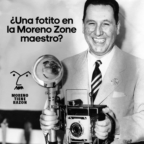 a man in a suit and tie is holding a camera in front of a sign that says moreno zone maestro
