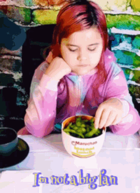 a girl with red hair is sitting at a table with a bowl of edamame beans
