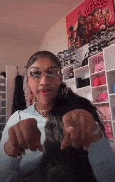 a girl wearing sunglasses is pointing at the camera in front of a wall that has a picture of a woman on it