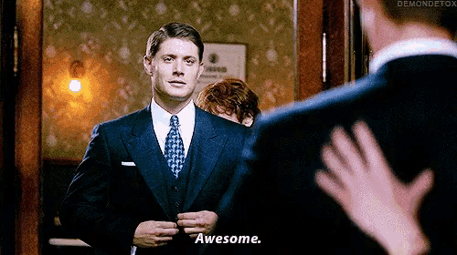 a man in a suit and tie is standing in front of a mirror and says awesome .