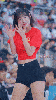 a girl in a red crop top and black shorts is waving at the camera