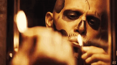 a man with a skull painted on his face is smoking a cigarette in front of a mirror .
