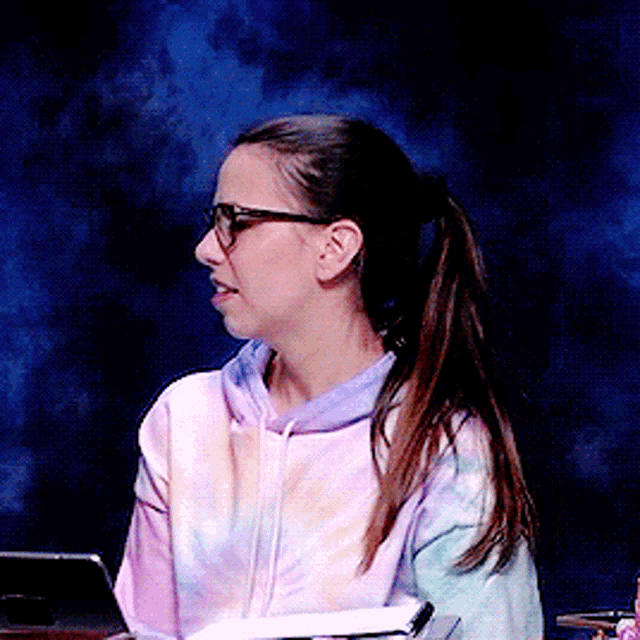 a woman wearing glasses and a tie dye sweatshirt sits in front of a laptop