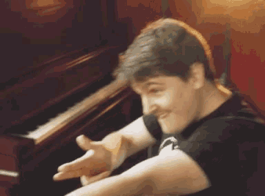 a man in a black shirt is sitting in front of a piano and making a funny face .