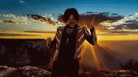 a man in a striped jacket stands on top of a rocky hill at sunset
