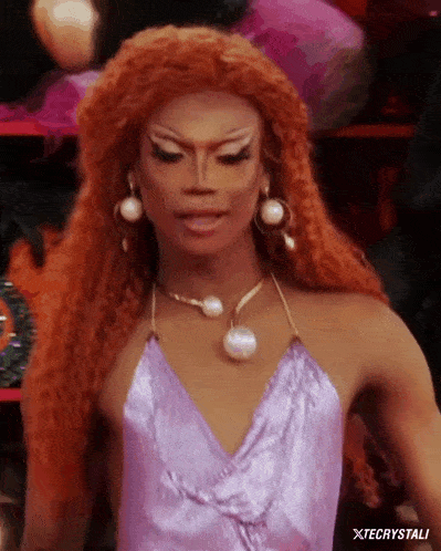 a drag queen with red hair is wearing a purple top and pearl earrings .