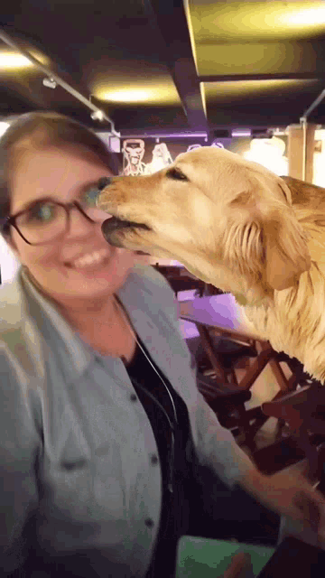 a woman with glasses is standing next to a dog that is licking her face