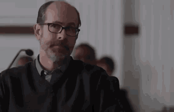 a man with glasses and a beard is sitting in a courtroom .