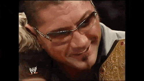 a man wearing glasses is holding a wrestling belt and smiling .