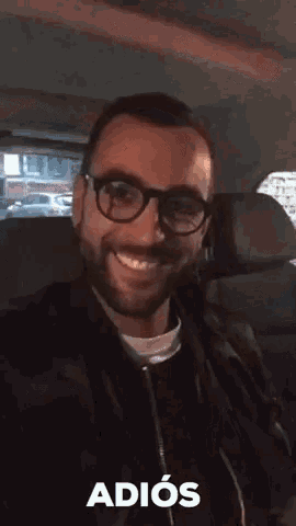 a man with glasses and a beard is sitting in the back seat of a car and says adios .