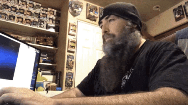 a man with a beard is typing on a laptop in front of a wall full of funko pop figures