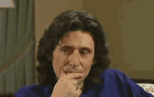 a man with long hair and a ring on his finger is sitting on a couch with his hand on his chin .