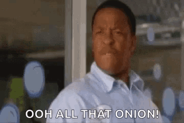 a man in a blue shirt is standing in front of a window and saying `` ooh all that onion '' .