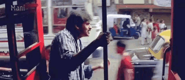 a man in a plaid shirt is standing in front of a red bus with mani written on it
