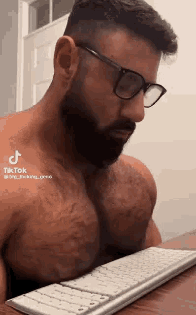 a shirtless man with a beard and glasses is sitting at a table using a keyboard .