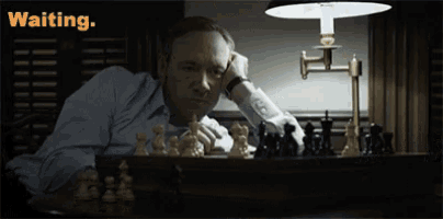 a man is sitting at a table with chess pieces and the word waiting is above him