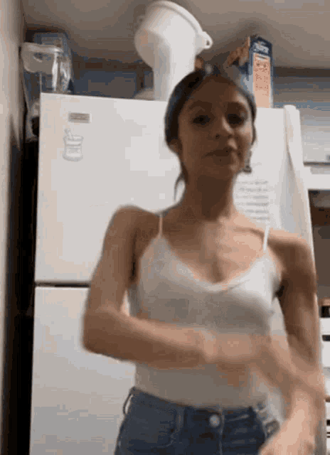 a woman in a white tank top is standing in front of a refrigerator in a kitchen .