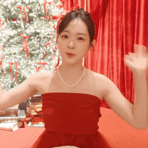 a woman in a red dress with a pearl necklace is waving in front of a christmas tree