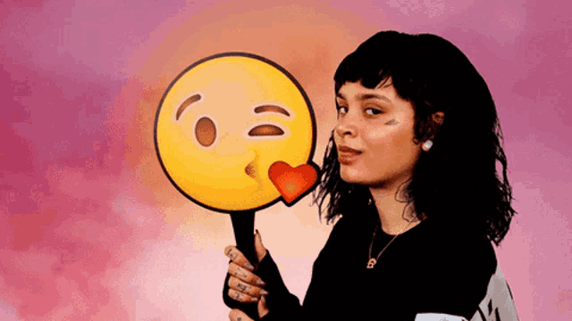 a woman with a tattoo on her face is holding a smiley face with a heart on it