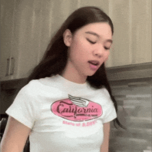 a woman wearing a white california t-shirt is standing in a kitchen