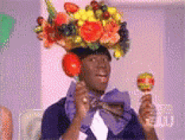 a man wearing a hat made of fruit and flowers is holding a lollipop .